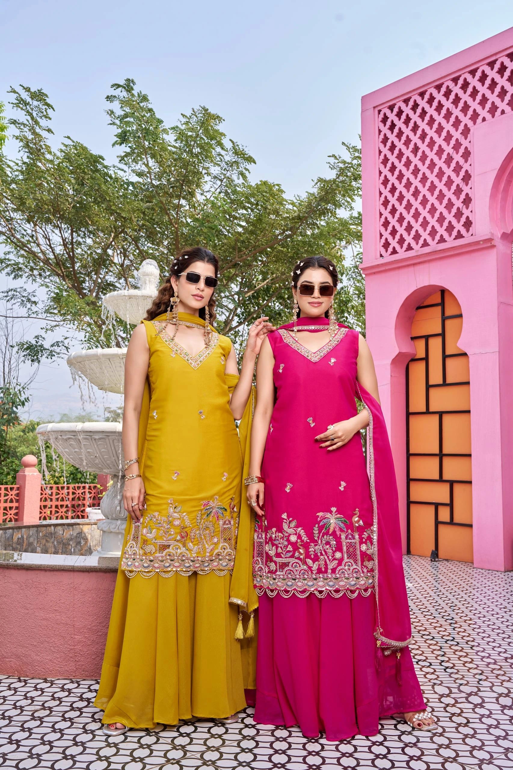 Two women in traditional yellow and pink dresses standing in front of a pink architectural structure.